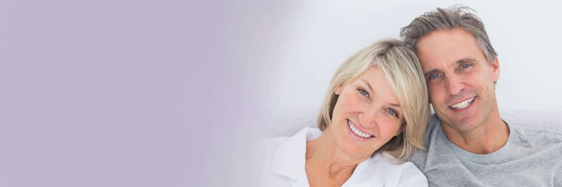 Smiling middle-aged couple posing together against a soft purple and white background.