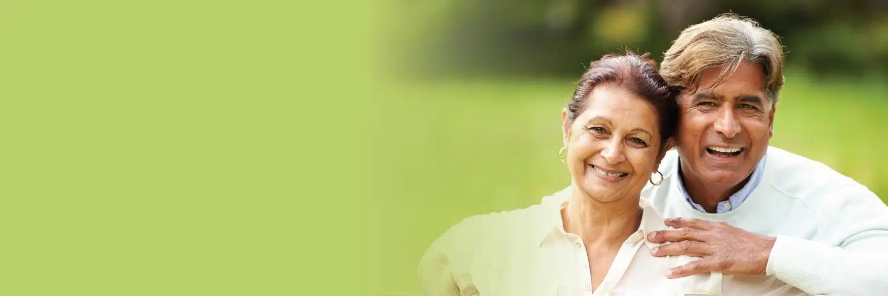 Smiling older couple outdoors in green setting, man's arm around woman's shoulder.