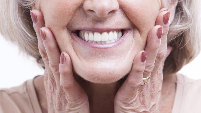 Smiling older woman touching her face, displaying healthy teeth and skin in dental care context.