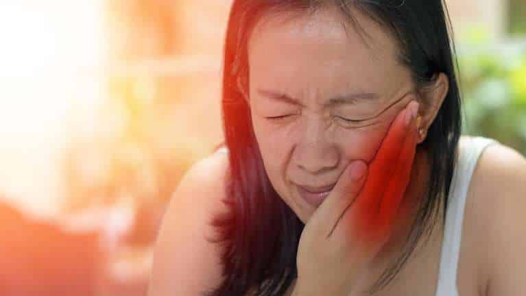 Woman holding cheek in pain with warm, bright outdoor lighting background.