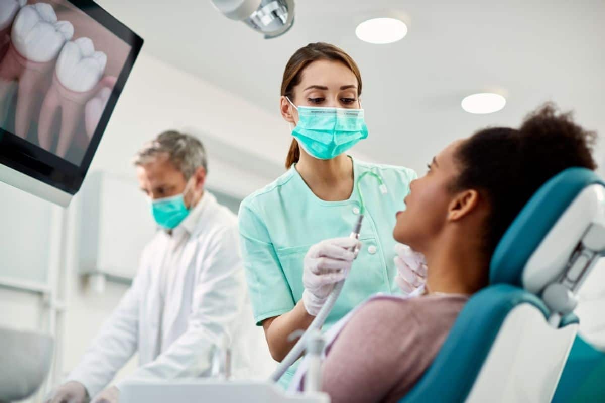 Dental hygienist in mask examines patient in dental chair with dentist observing in background.