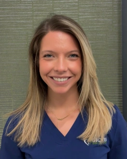 Professional headshot of smiling woman in dark blue scrubs against neutral dental office background.